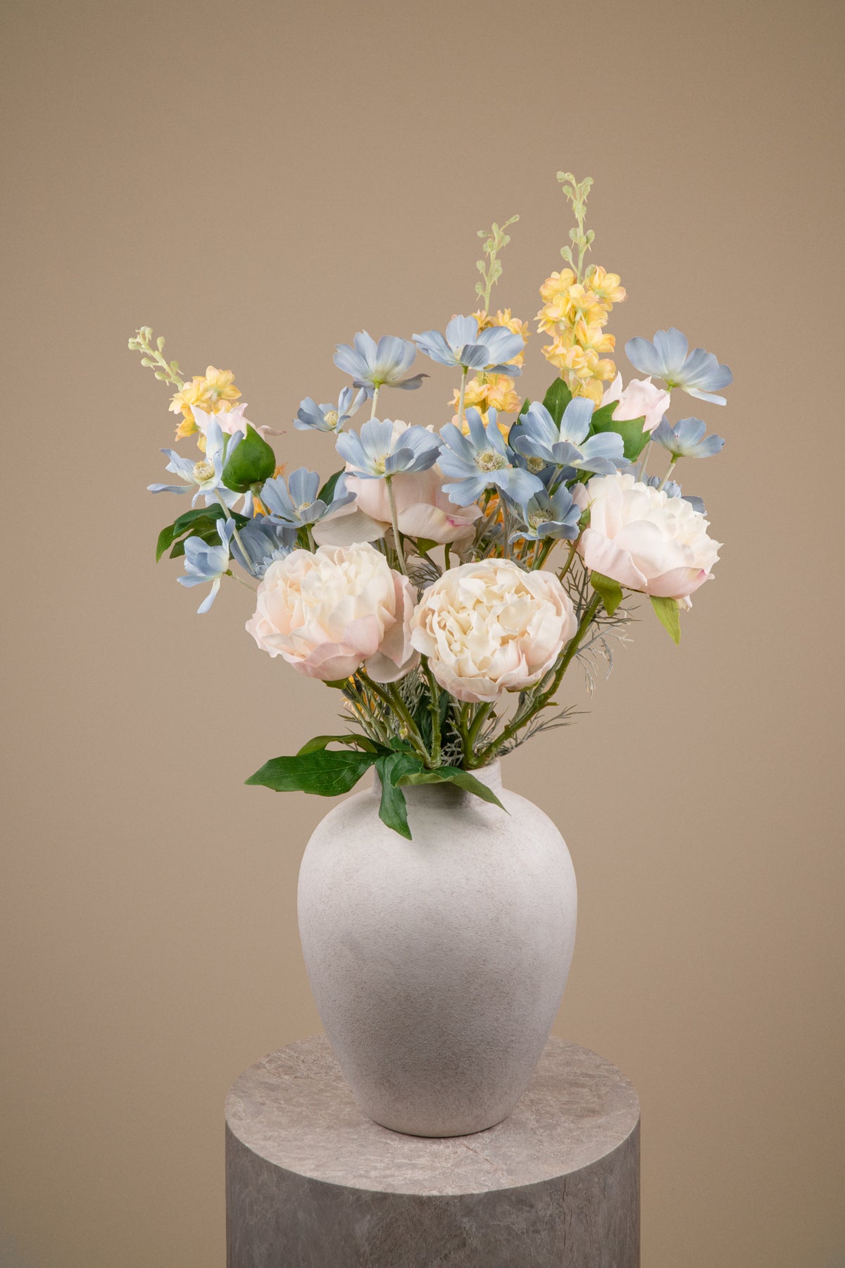 An artificial floral arrangement featuring light blue hydrangeas, pale pink peonies, and yellow flowers with green foliage, displayed in a matte gray ceramic vase on a round stone pedestal against a solid beige background.
