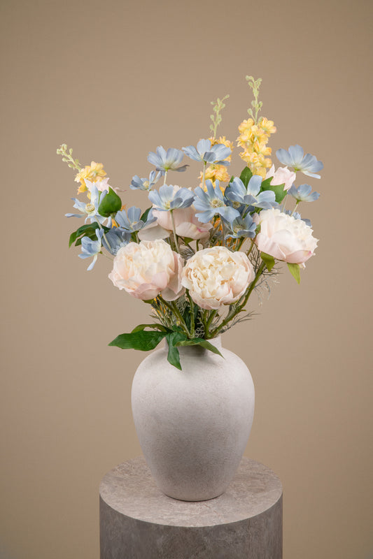An artificial floral arrangement featuring light blue hydrangeas, pale pink peonies, and yellow flowers with green foliage, displayed in a matte gray ceramic vase on a round stone pedestal against a solid beige background.