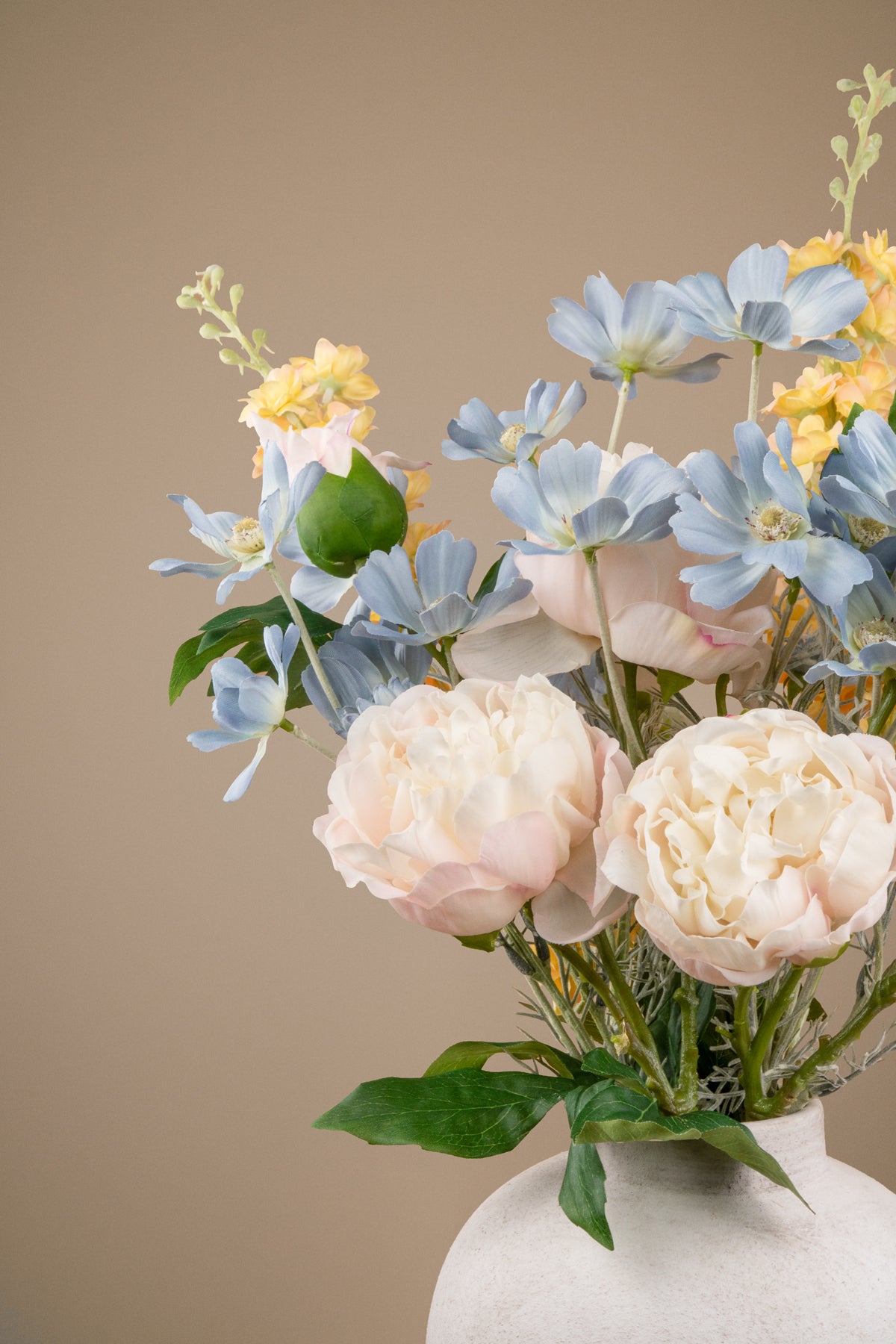 A floral arrangement featuring pale blush peonies, light blue anemone-like flowers, yellow blossoms, and green leaves in a white ceramic vase against a solid warm beige background.