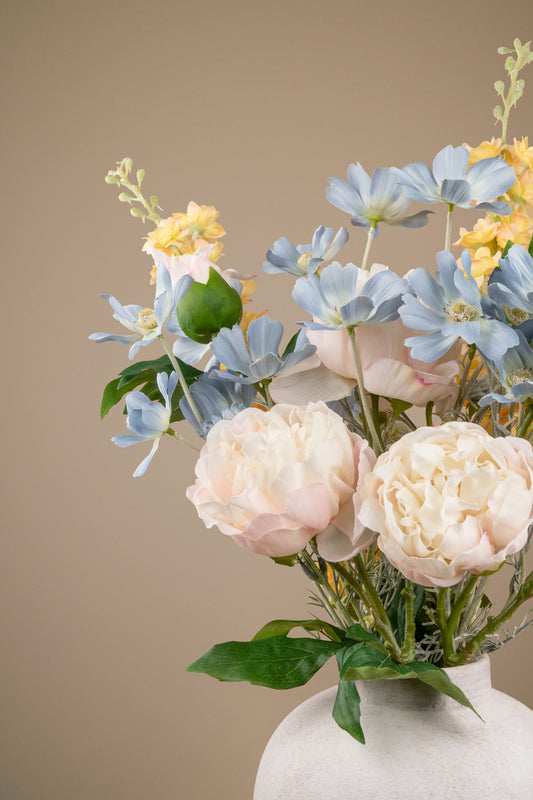A floral arrangement featuring pale blush peonies, light blue anemone-like flowers, yellow blossoms, and green leaves in a white ceramic vase against a solid warm beige background.