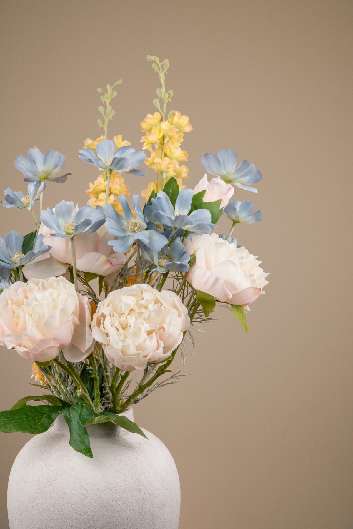 A floral arrangement featuring pale blush peonies, light blue flowers, and yellow spikes with green foliage, presented in a matte white ceramic vase against a solid warm beige background.