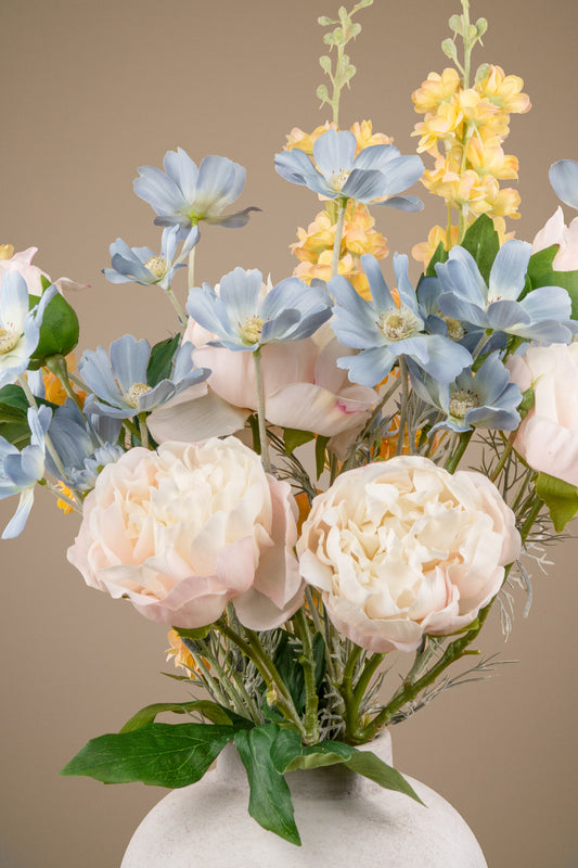 An artificial floral arrangement featuring pale pink peonies, light blue flowers, yellow delphiniums, and green foliage, displayed in a textured white ceramic vase against a solid warm beige background.