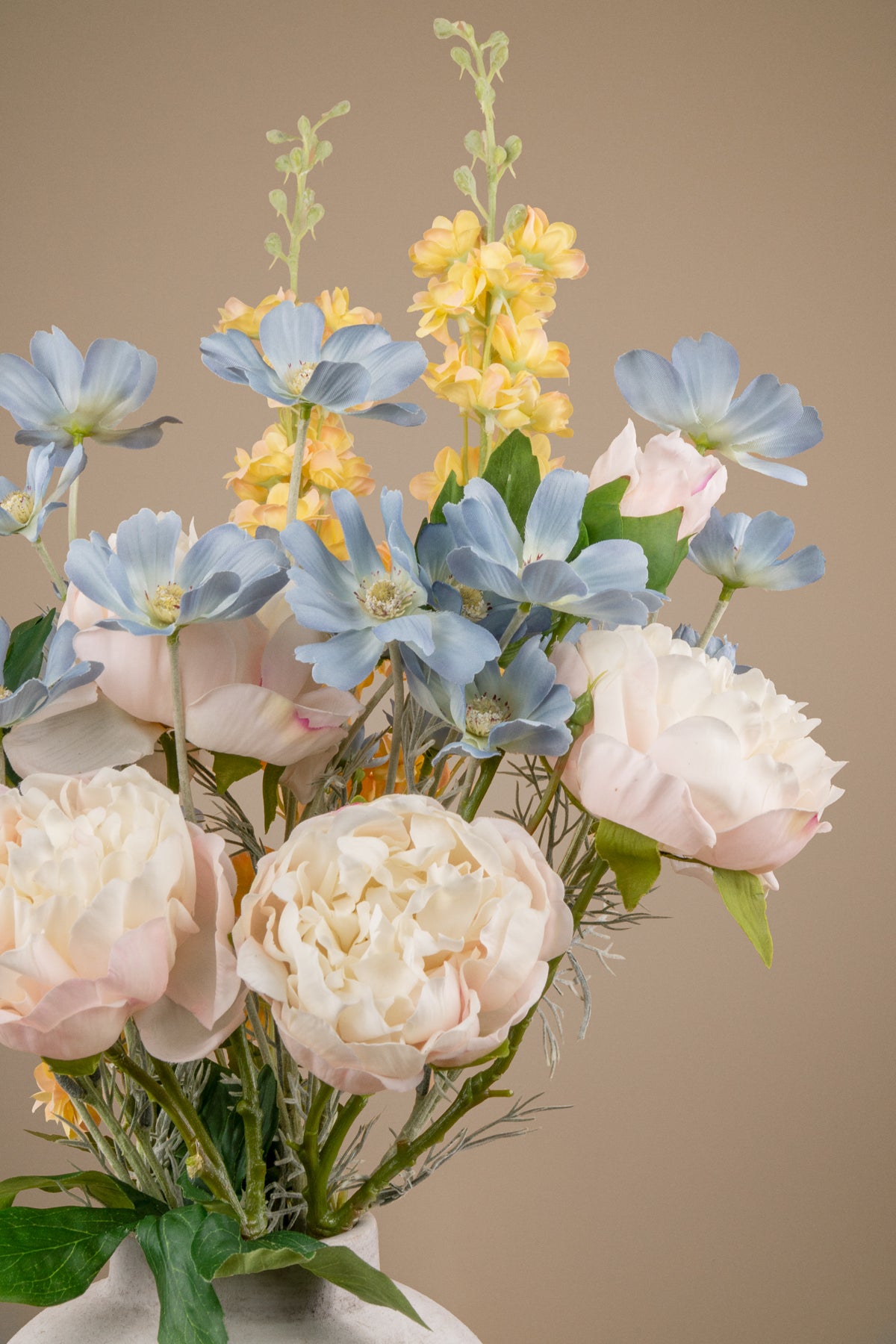 A bouquet of artificial flowers in a white vase, featuring pale pink and cream peonies, light blue flowers, and yellow spikes, set against a solid beige background.