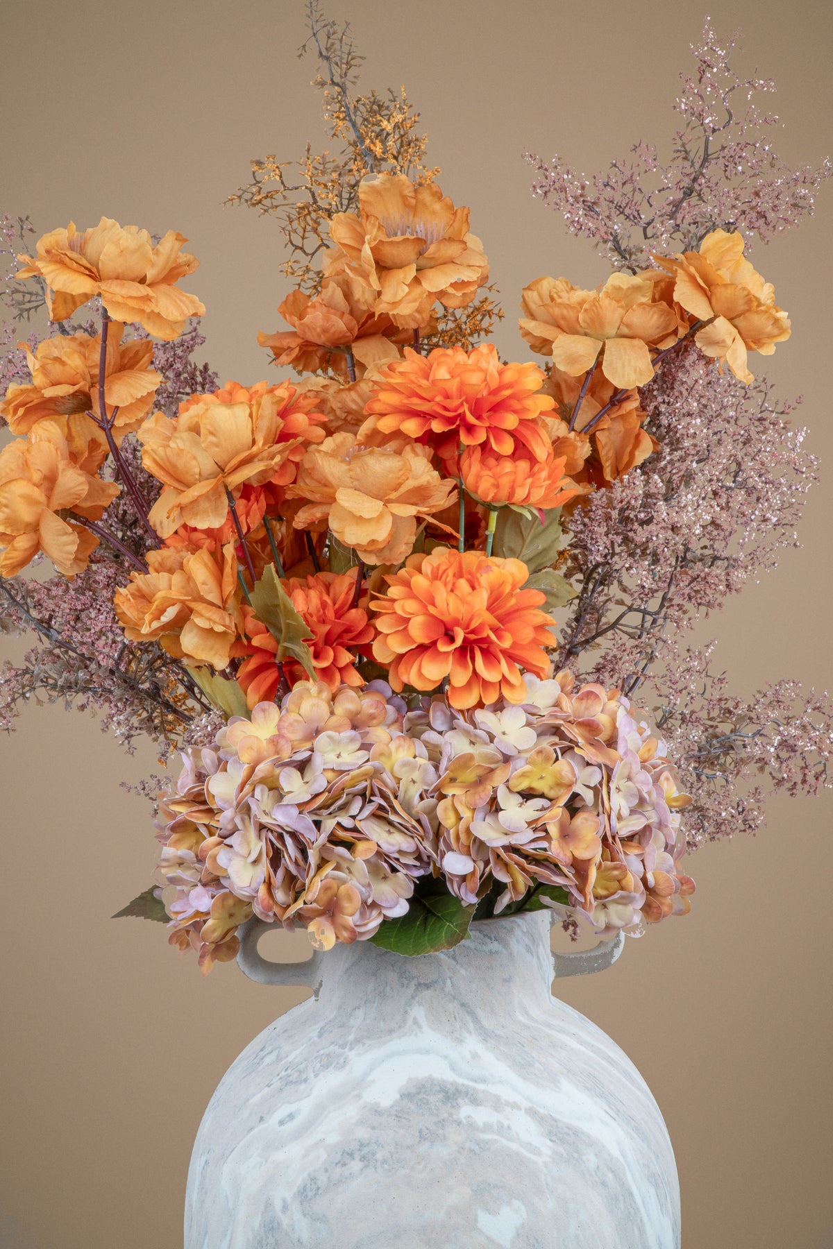An artificial floral arrangement featuring orange and peach dahlias, peach hydrangeas, and dusty lavender filler stems in a distressed white ceramic vase against a solid beige background.