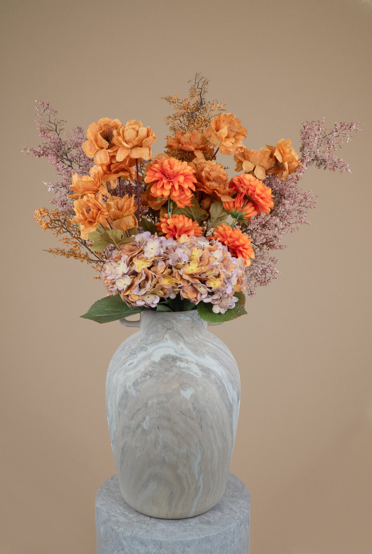 An artificial floral arrangement featuring orange and peach flowers, light purple filler stems, and green leaves, displayed in a gray marble-patterned ceramic vase on a matching gray pedestal against a solid beige background.