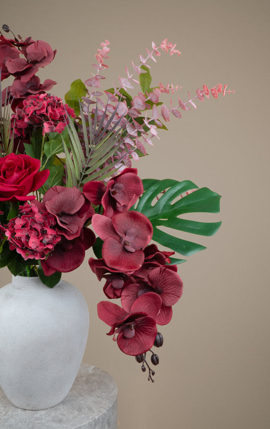 A floral arrangement in a matte white ceramic vase, featuring deep burgundy orchids, pink hydrangeas, green monstera leaves, and delicate light pink sprigs against a solid beige background.