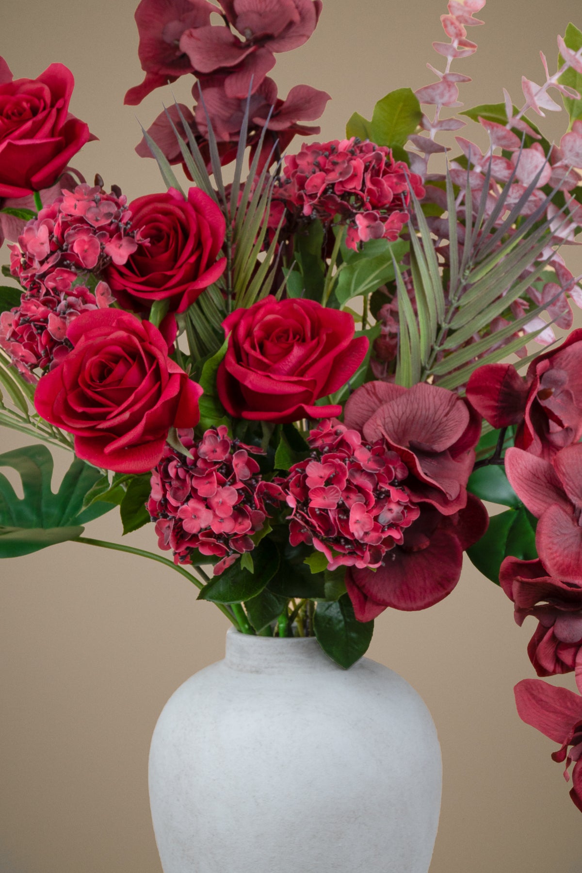 A close-up of a floral arrangement featuring deep red roses, burgundy hydrangeas, and green foliage in a matte white ceramic vase against a solid beige background.