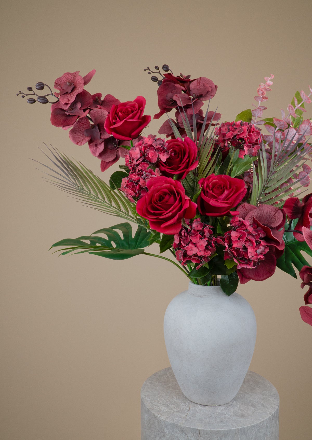 A floral arrangement featuring deep red roses, burgundy hydrangeas, pink blossoms, and green foliage in a matte white ceramic vase, displayed on a stone pedestal against a warm beige background.