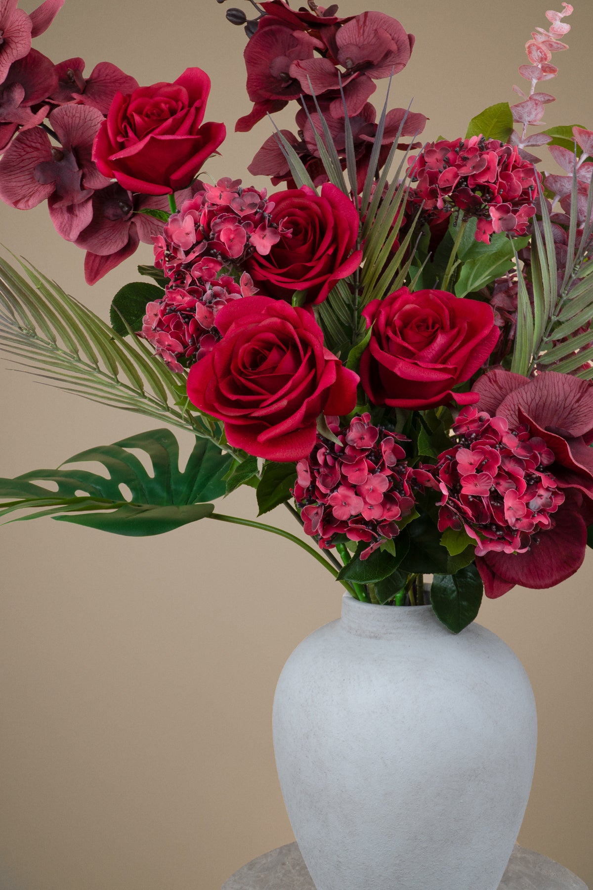 A floral arrangement featuring deep red roses, burgundy hydrangeas, and green foliage in a matte white ceramic vase against a solid beige background.