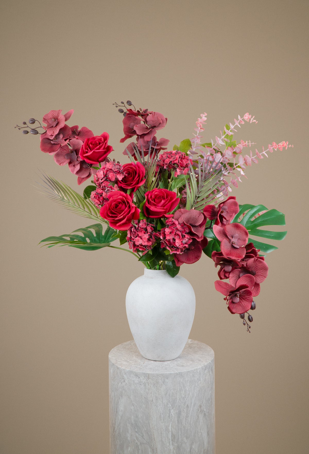 A floral arrangement featuring deep pink roses, burgundy orchids, green foliage, and light pink filler stems in a matte white ceramic vase, displayed on a light gray stone pedestal against a solid beige background.