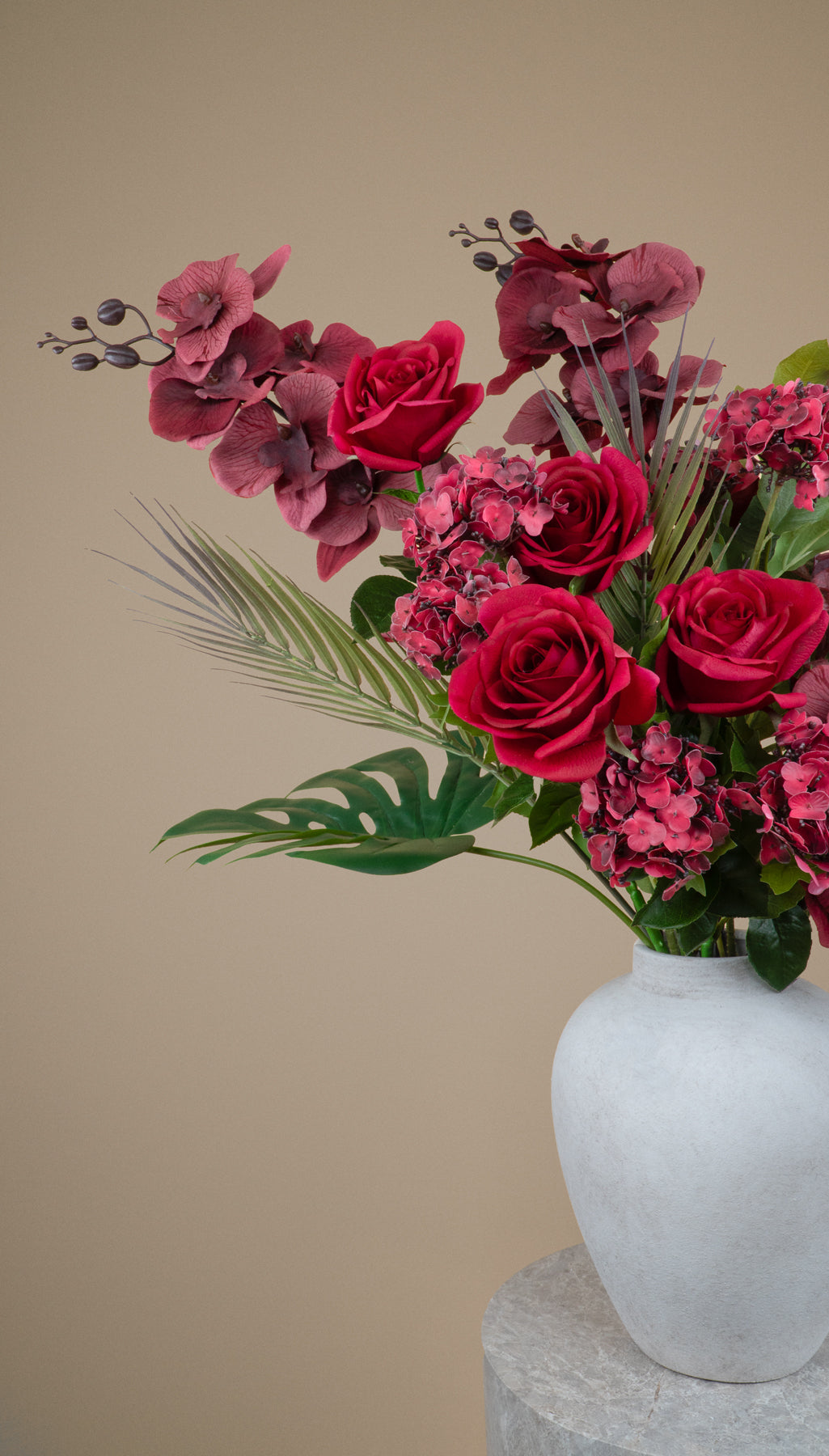 A floral arrangement featuring deep red roses, burgundy hydrangeas, and green foliage in a white ceramic vase, set against a solid beige background.