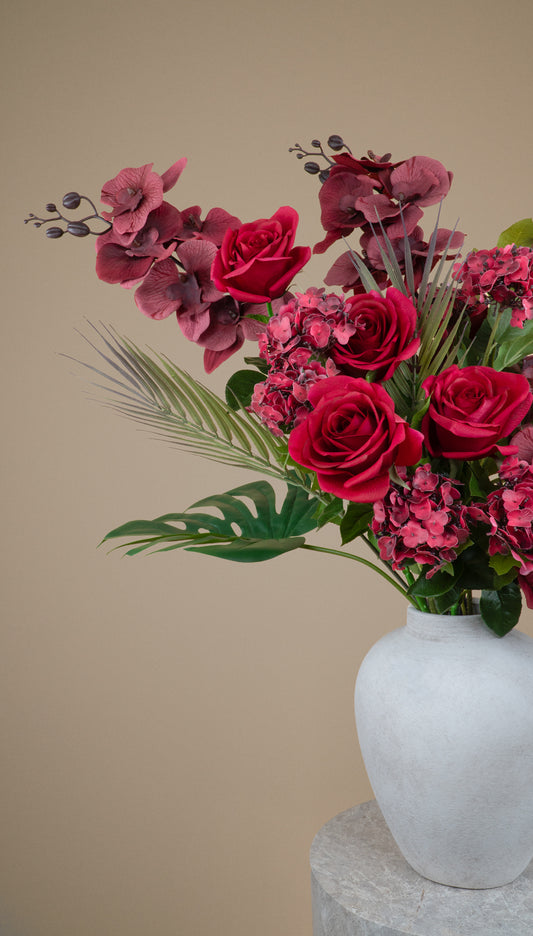 A floral arrangement featuring deep red roses, burgundy hydrangeas, and green foliage in a white ceramic vase, set against a solid beige background.
