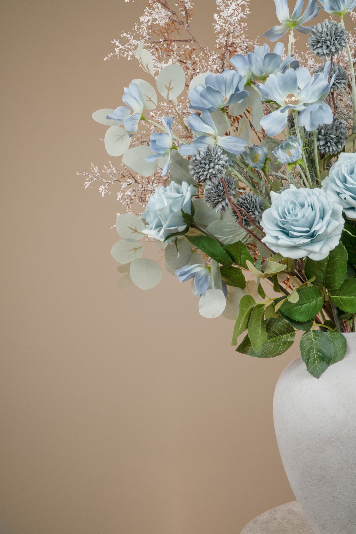 A floral arrangement featuring light blue roses, dusty blue kikus, baby blue cosmos chrysanthemums, and delicate white filler stems and white branches in a white ceramic vase against a solid beige background.