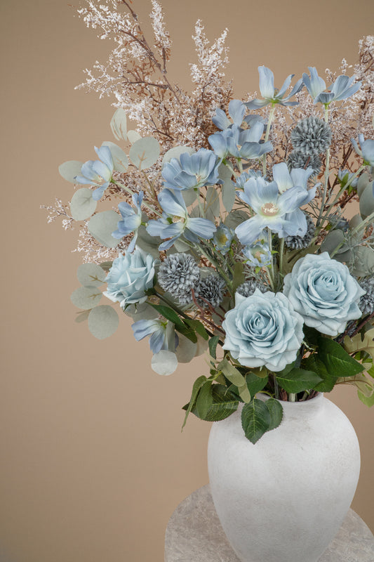 A close-up of a floral arrangement featuring light blue roses, dusty blue kikus, baby blue cosmos chrysanthemums, and delicate white filler stems and white branches in a textured white ceramic vase, set against a solid beige background.
