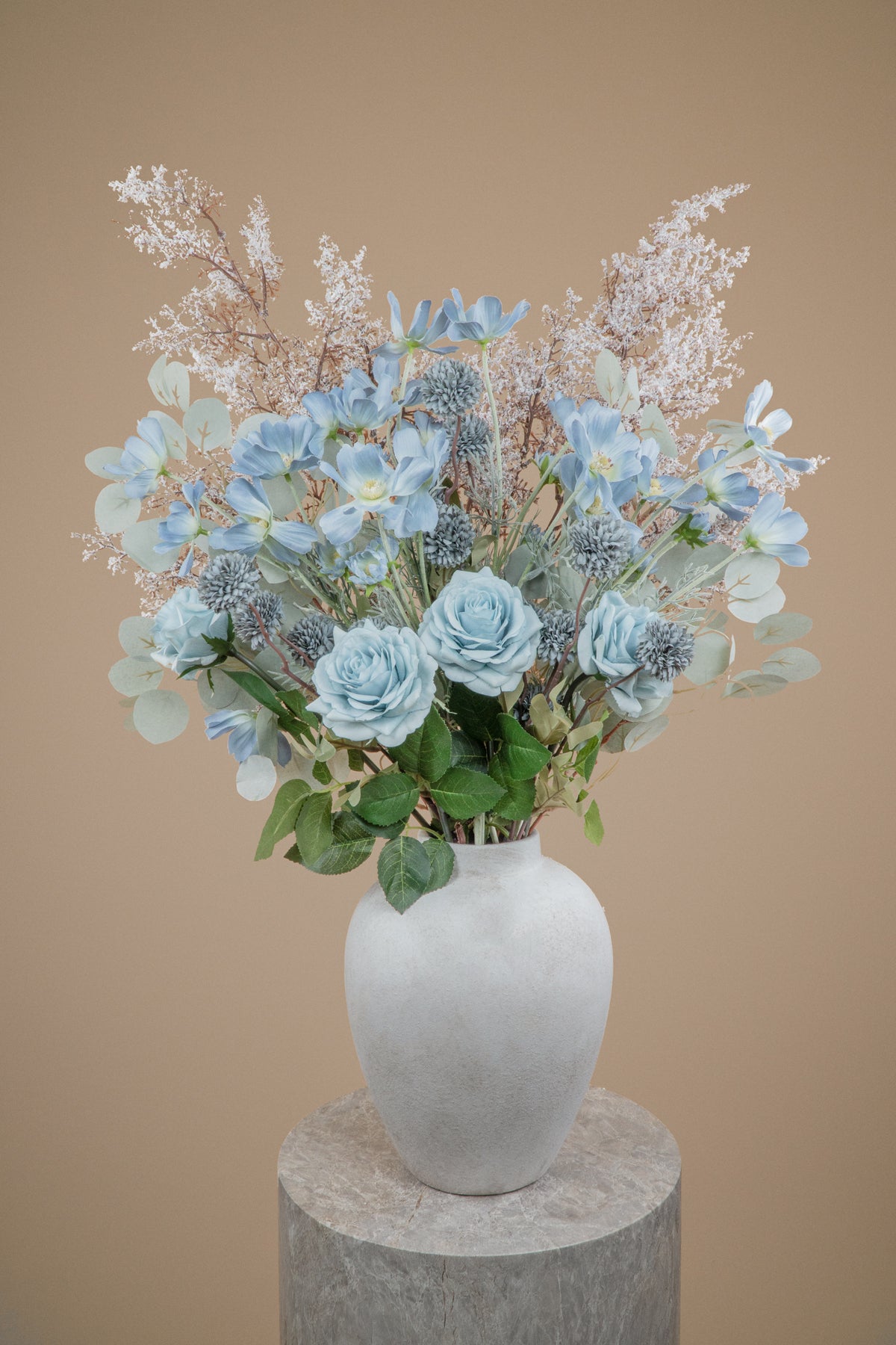 A decorative faux floral arrangement with light blue roses, dusty blue kikus, baby blue cosmos chrysanthemums, and delicate white filler stems and white branches in a matte white ceramic vase, displayed on a round stone pedestal against a warm beige background.
