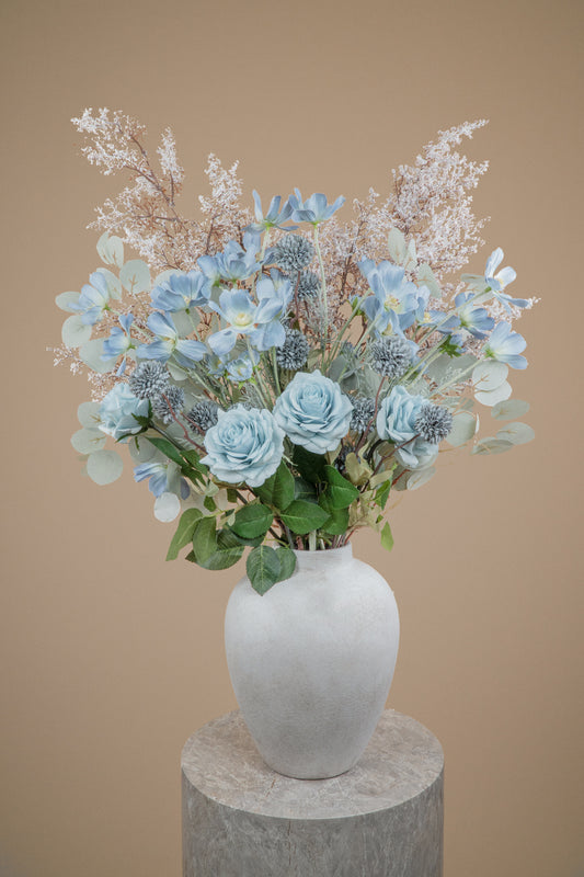 A decorative faux floral arrangement with light blue roses, dusty blue kikus, baby blue cosmos chrysanthemums, and delicate white filler stems and white branches in a matte white ceramic vase, displayed on a round stone pedestal against a warm beige background.