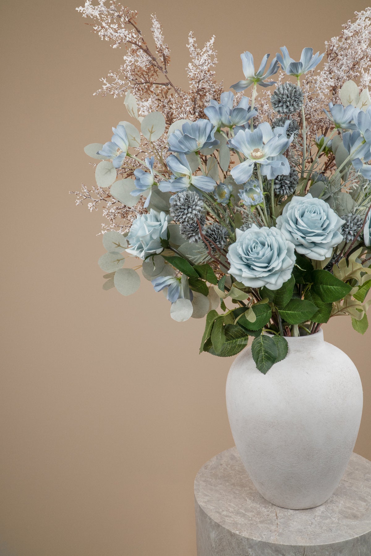 A floral arrangement featuring light blue roses, dusty blue kikus, baby blue cosmos chrysanthemums, and delicate white filler stems and white branches in a textured white ceramic vase, set against a solid beige background on a stone pedestal.