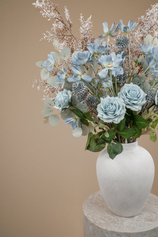 A floral arrangement featuring light blue roses, dusty blue kikus, baby blue cosmos chrysanthemums, and delicate white filler stems and white branches in a textured white ceramic vase, set against a solid beige background on a stone pedestal.