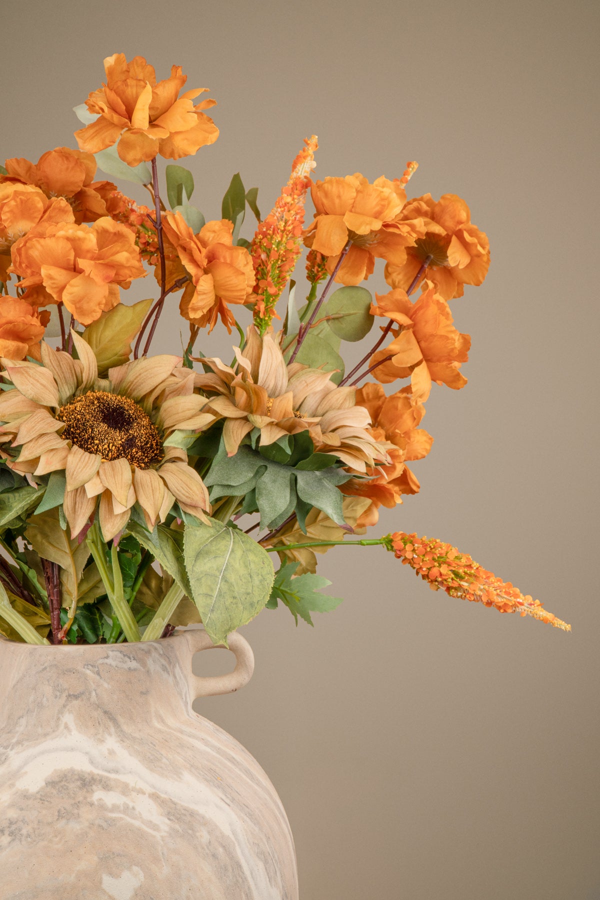 An artificial autumn floral arrangement in a white marbled ceramic vase, featuring orange roses, sunflowers, and green foliage against a solid taupe background.