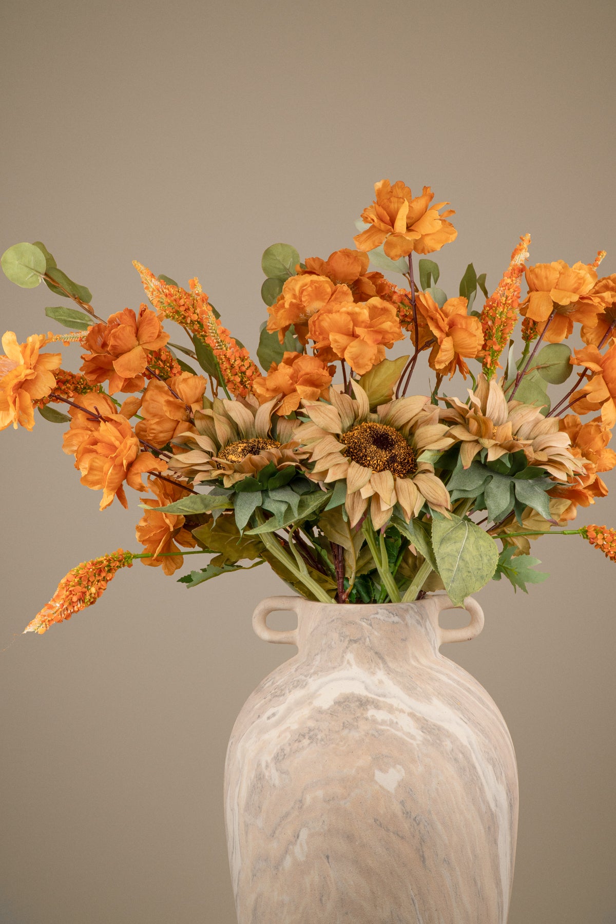 An artificial autumn floral arrangement featuring orange flowers, tan sunflowers, and green leaves, displayed in a textured beige ceramic vase against a solid taupe background.