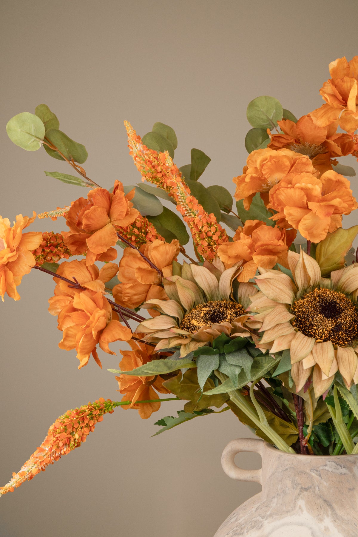 An autumn floral arrangement featuring orange peony-like flowers, tan sunflowers, green leaves, and orange seed pods in a white ceramic vase against a solid taupe background.