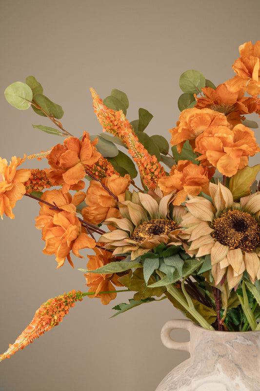 An autumn floral arrangement featuring orange peony-like flowers, tan sunflowers, green leaves, and orange seed pods in a white ceramic vase against a solid taupe background.