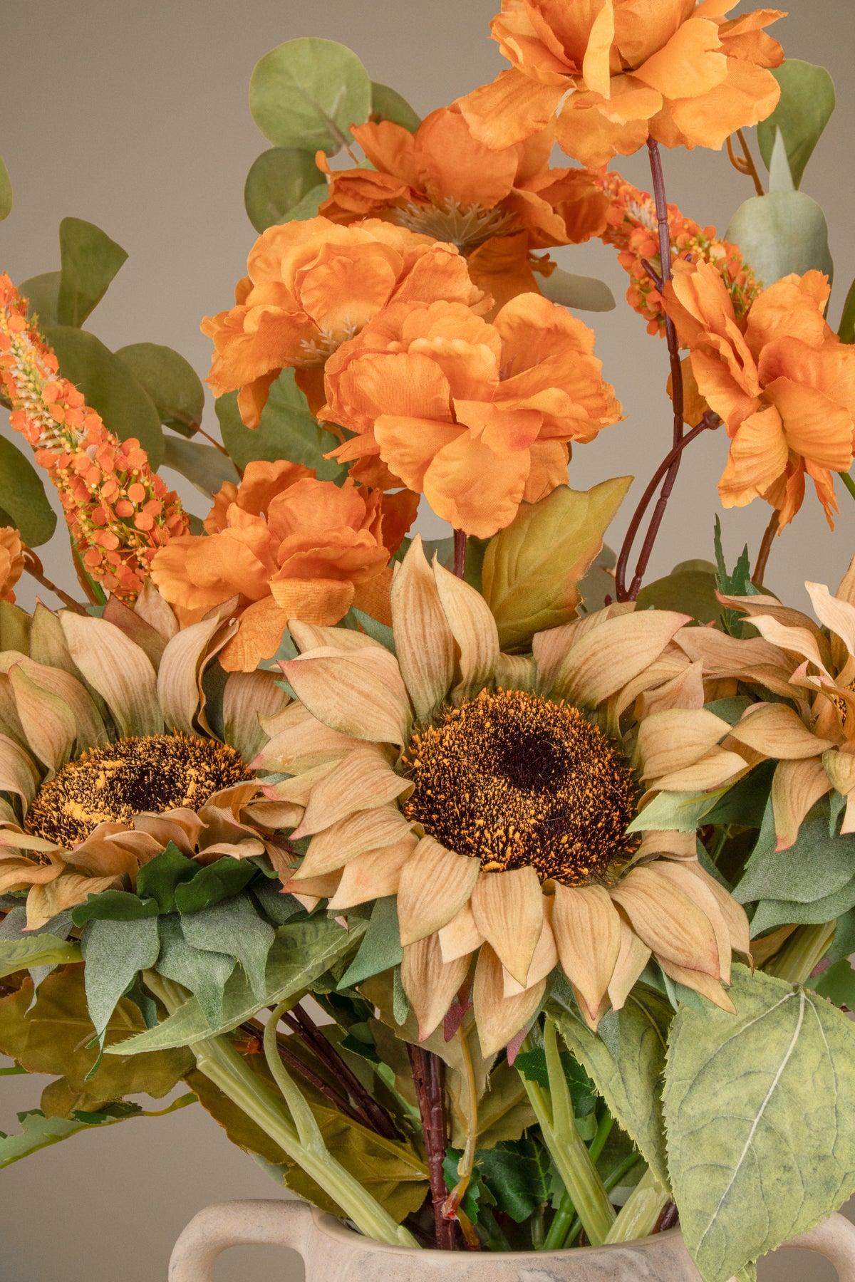 An autumnal floral arrangement featuring large sunflowers, orange peony-like blooms, green leaves, and orange coneflowers in a light-colored ceramic vase against a neutral beige background.