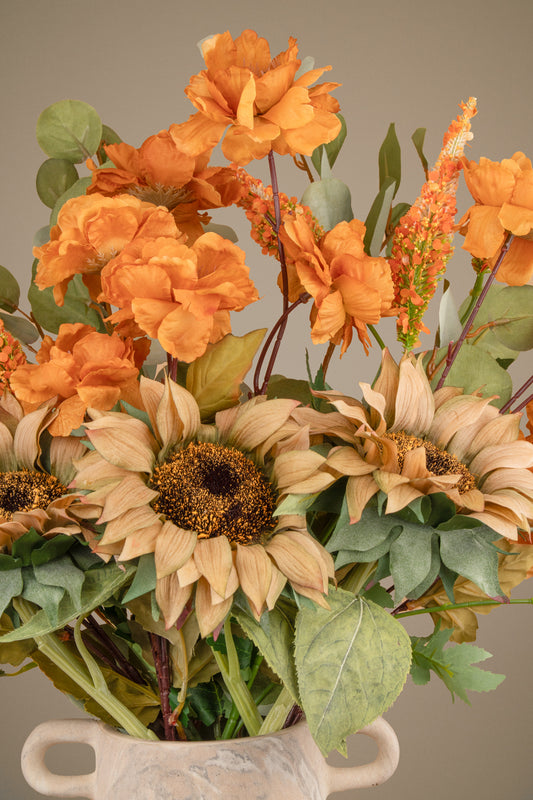 An artificial autumn floral arrangement featuring orange peony-like flowers, tan sunflowers, green leaves, and stems in a small white ceramic pot against a solid taupe background.