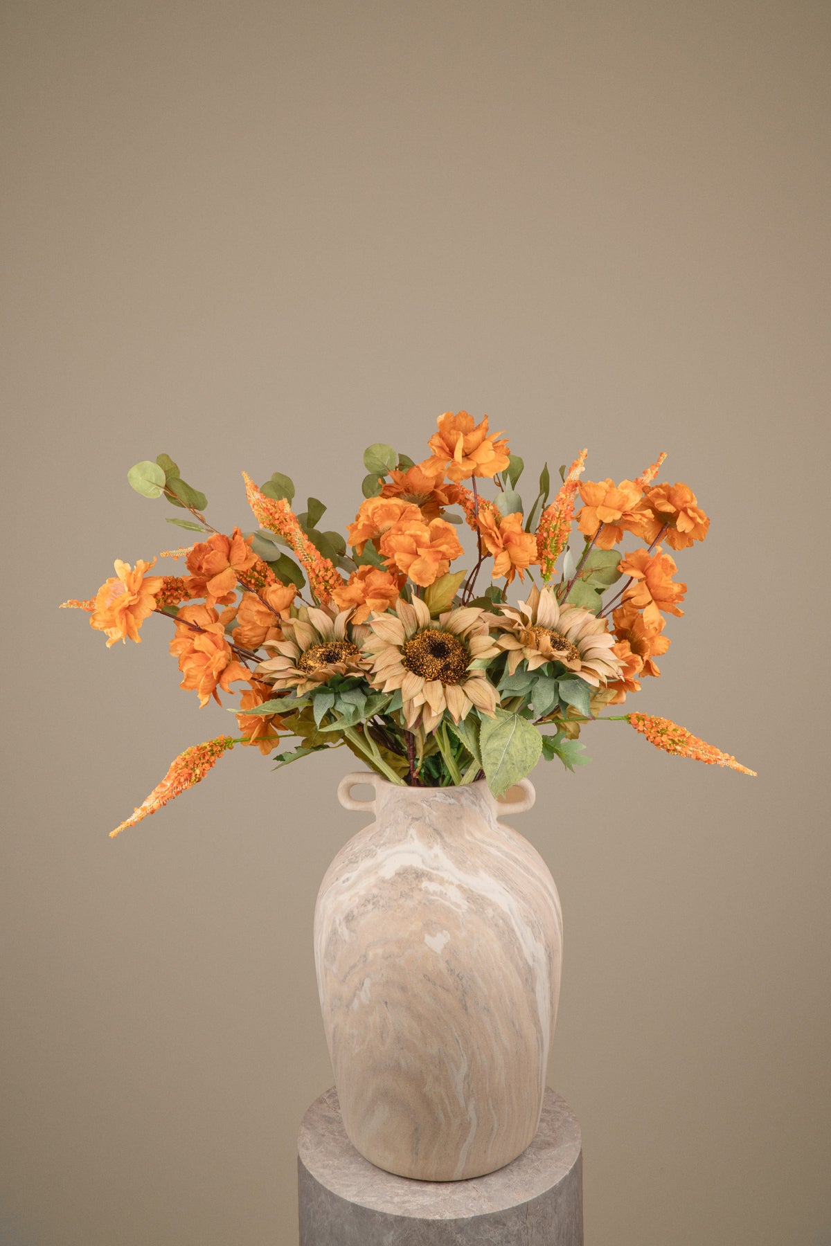 A faux autumn flower arrangement featuring orange blossoms, sunflowers, and green foliage in a cream-colored marble vase, displayed on a gray pedestal against a solid taupe background.