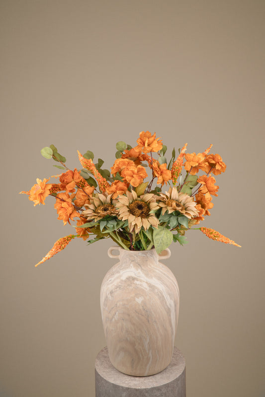 A faux autumn flower arrangement featuring orange blossoms, sunflowers, and green foliage in a cream-colored marble vase, displayed on a gray pedestal against a solid taupe background.