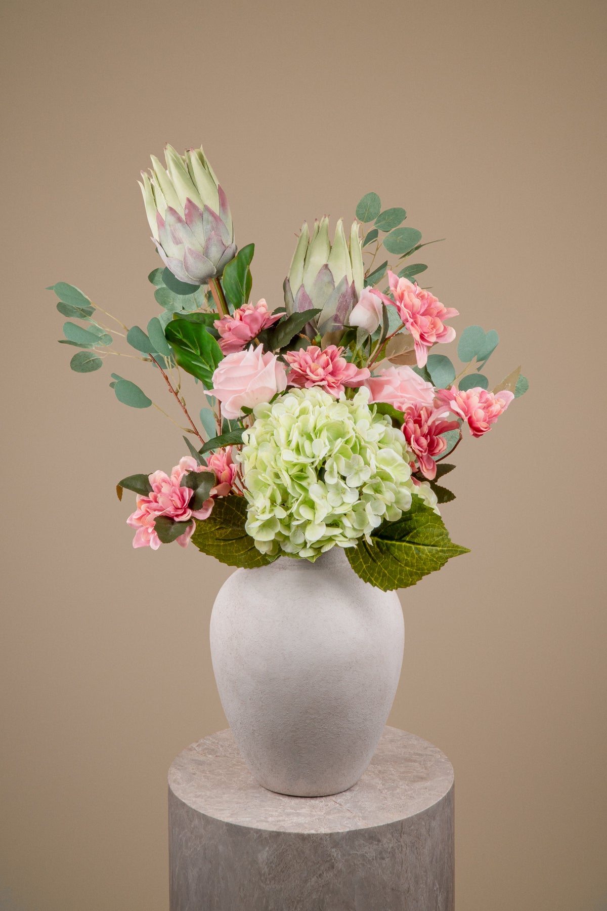 A floral arrangement featuring light green hydrangea, pink carnations, and protea flowers with green eucalyptus leaves, presented in a matte white ceramic vase on a round gray pedestal against a solid beige background.