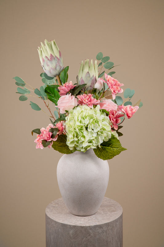 A floral arrangement featuring light green hydrangea, pink carnations, and protea flowers with green eucalyptus leaves, presented in a matte white ceramic vase on a round gray pedestal against a solid beige background.