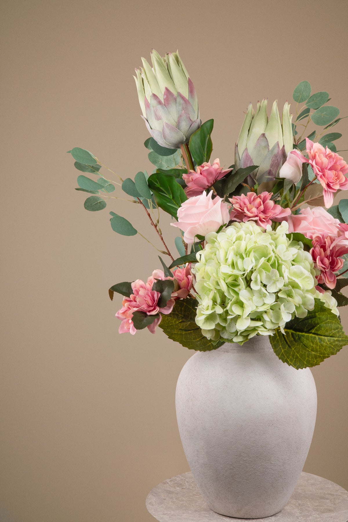 A floral arrangement featuring light green hydrangea, pink carnations, and protea flowers with green eucalyptus leaves, presented in a matte white ceramic vase on a light stone pedestal against a solid beige background.