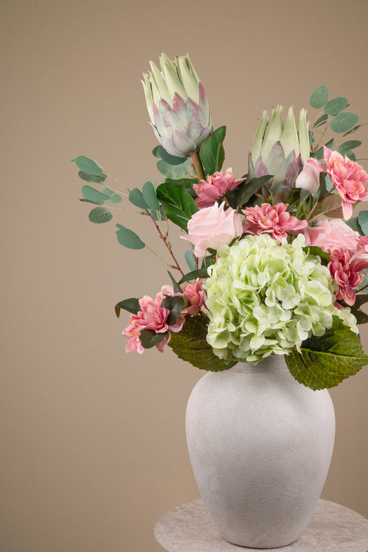 A floral arrangement featuring light green hydrangea, pink carnations, and protea flowers with green eucalyptus leaves, presented in a matte white ceramic vase on a light stone pedestal against a solid beige background.