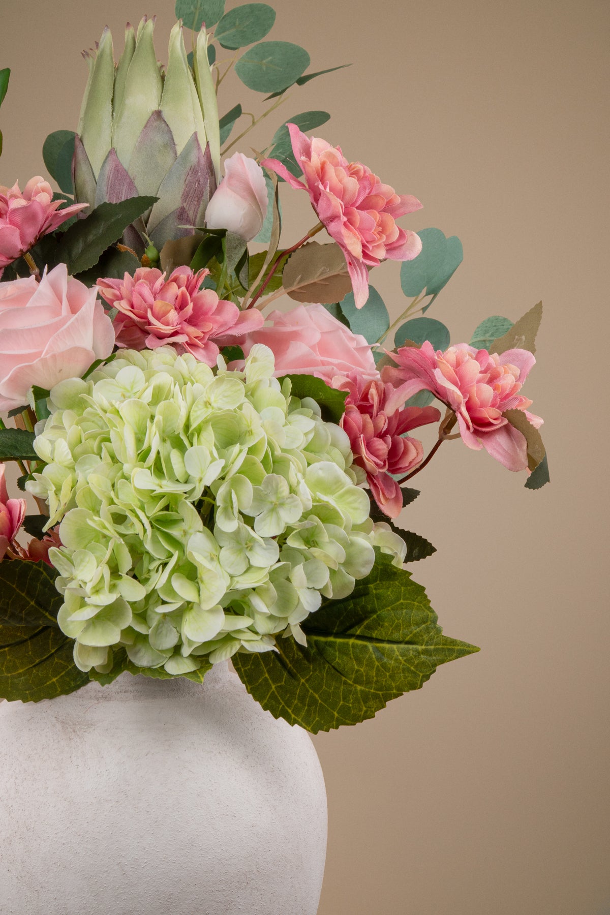 A floral arrangement featuring a large pale green hydrangea, pink roses, pink protea blooms, green eucalyptus leaves, and a green-and-white protea cone, displayed in a white textured vase against a warm beige background.