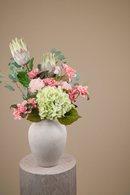 A floral arrangement featuring pink carnations, pale green hydrangea blooms, and protea flowers in a textured white ceramic vase, displayed on a round stone pedestal against a solid beige background.