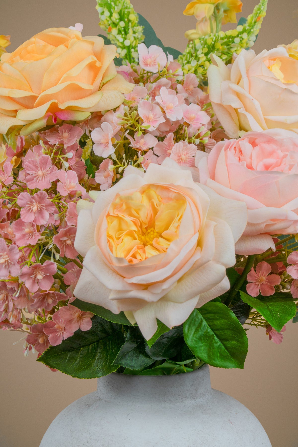 A close-up of a floral arrangement featuring peach and pale pink roses, small pink blossoms, green leaves, and sprigs of greenery, presented in a white ceramic vase against a warm beige background.