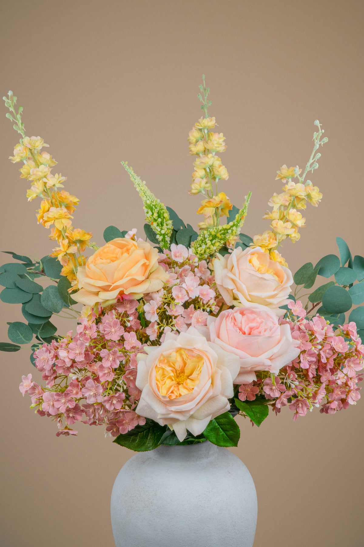 A floral arrangement featuring peach and blush roses, pink hydrangeas, yellow snapdragons, and green eucalyptus leaves in a round white ceramic vase against a solid beige background.