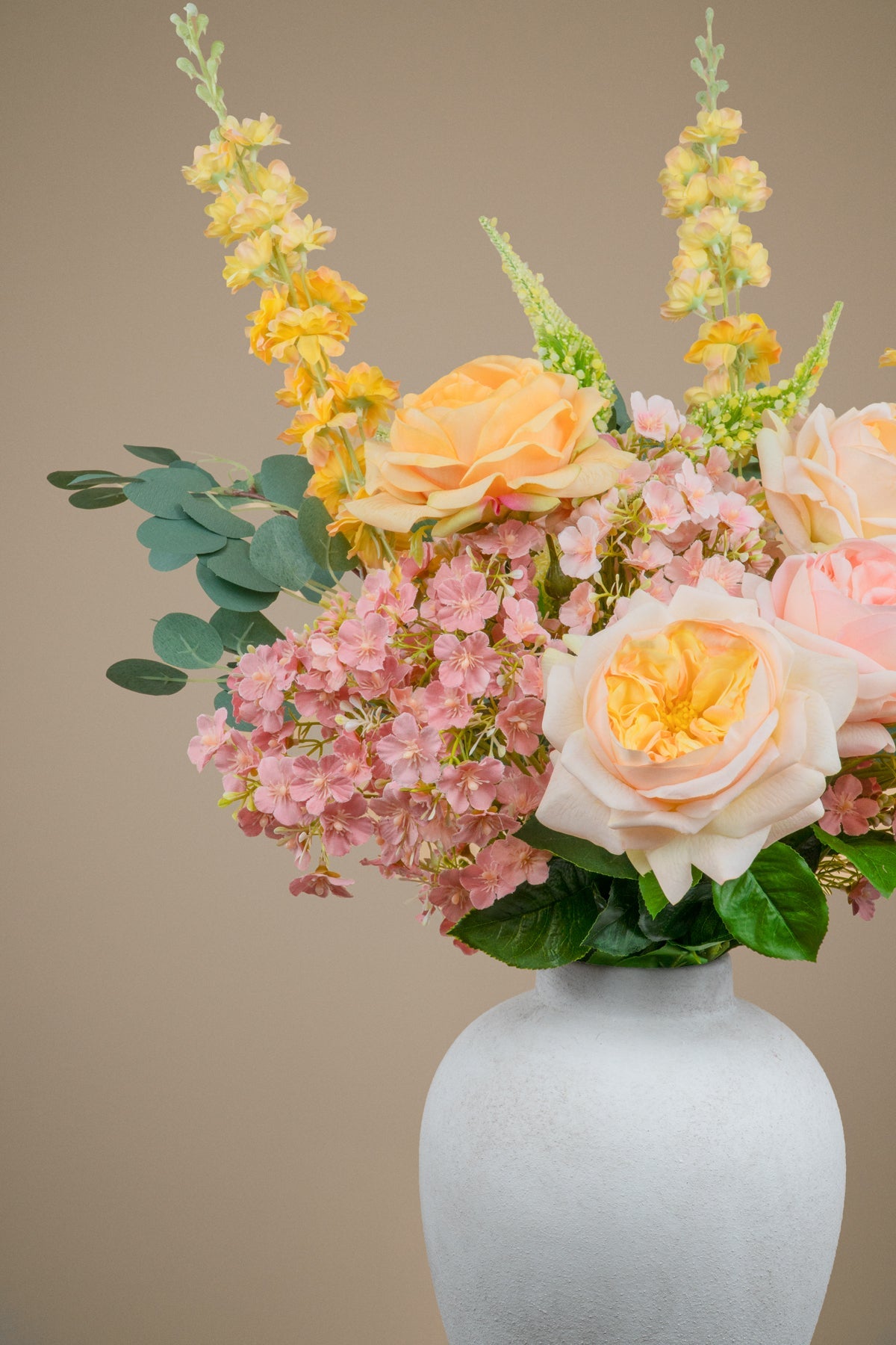 A floral arrangement featuring peach roses, light pink hydrangeas, yellow snapdragons, and green eucalyptus leaves in a textured white ceramic vase against a solid beige background.