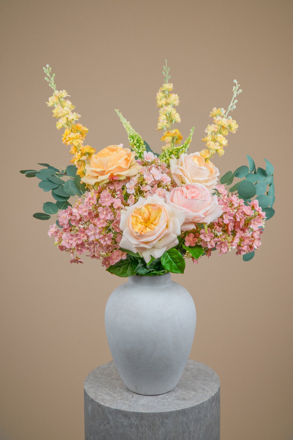A floral arrangement featuring pink hydrangeas, peach and blush roses, yellow snapdragons, and green eucalyptus leaves in a matte light gray ceramic vase, displayed on a round gray pedestal against a solid beige background.