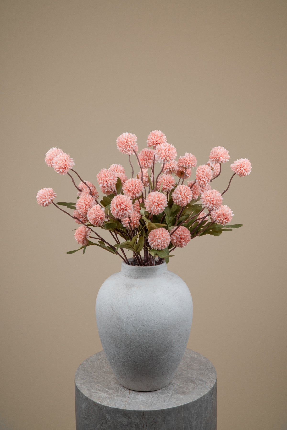 A bouquet of pink globe flowers with green leaves arranged in a matte light gray ceramic vase, set on a round gray pedestal against a solid warm beige background.