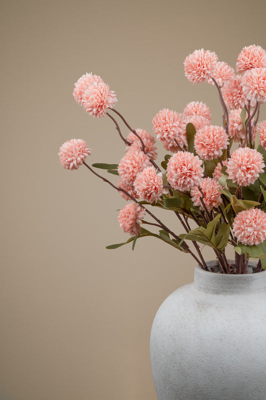 Pink pom-pom dahlia artificial flowers arranged in a textured light gray ceramic vase against a solid beige background.
