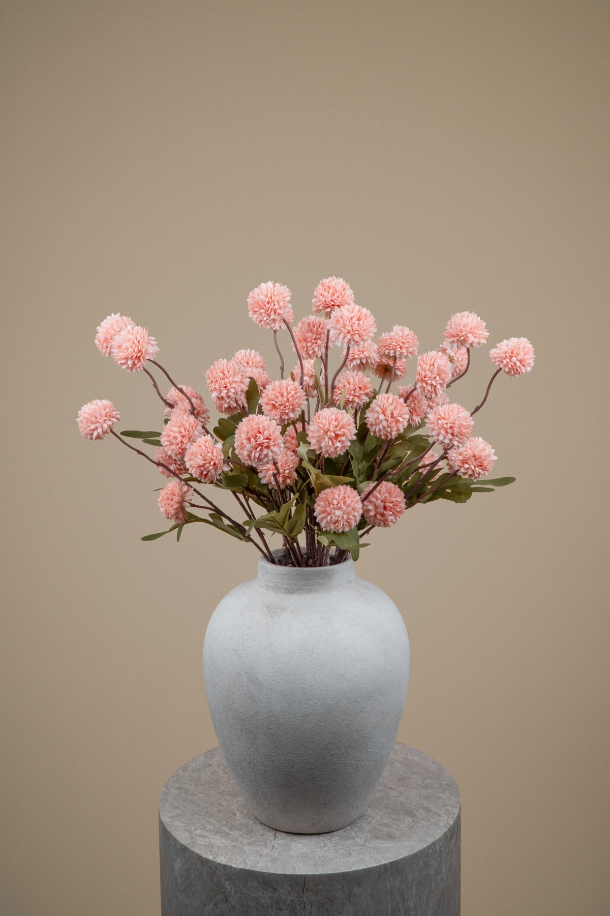Pink globe flowers arranged in a matte light gray ceramic vase, set on a round gray pedestal against a warm beige background.