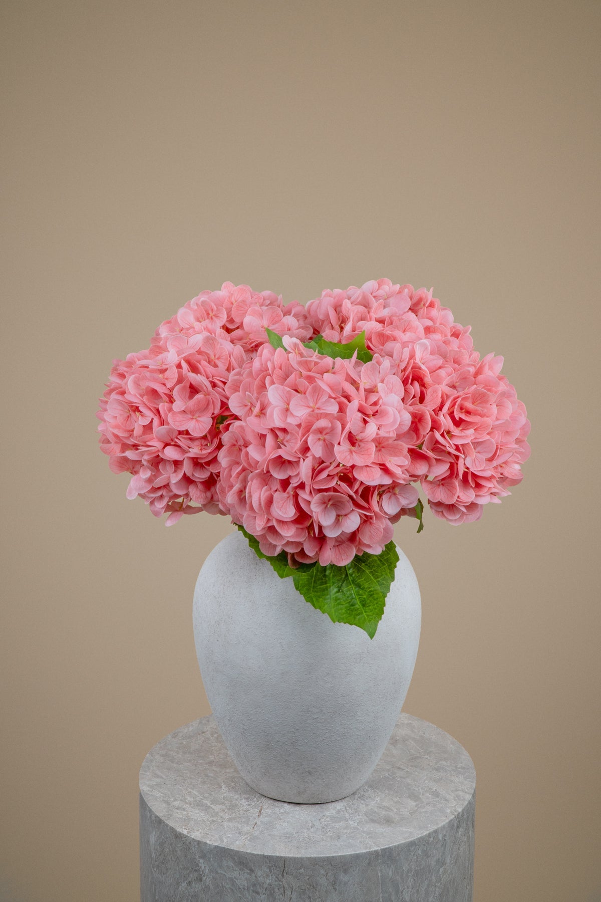 Three pink hydrangea flower bouquets with green leaves arranged in a white ceramic vase, set on a round gray stone pedestal against a solid beige background.