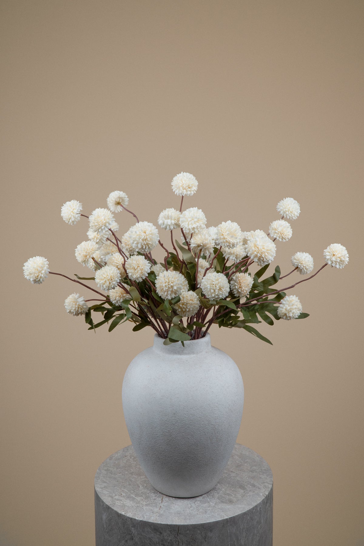 White pom-pom dahlia artificial flowers arranged in a matte light gray ceramic vase, displayed on a round gray pedestal against a solid beige background.