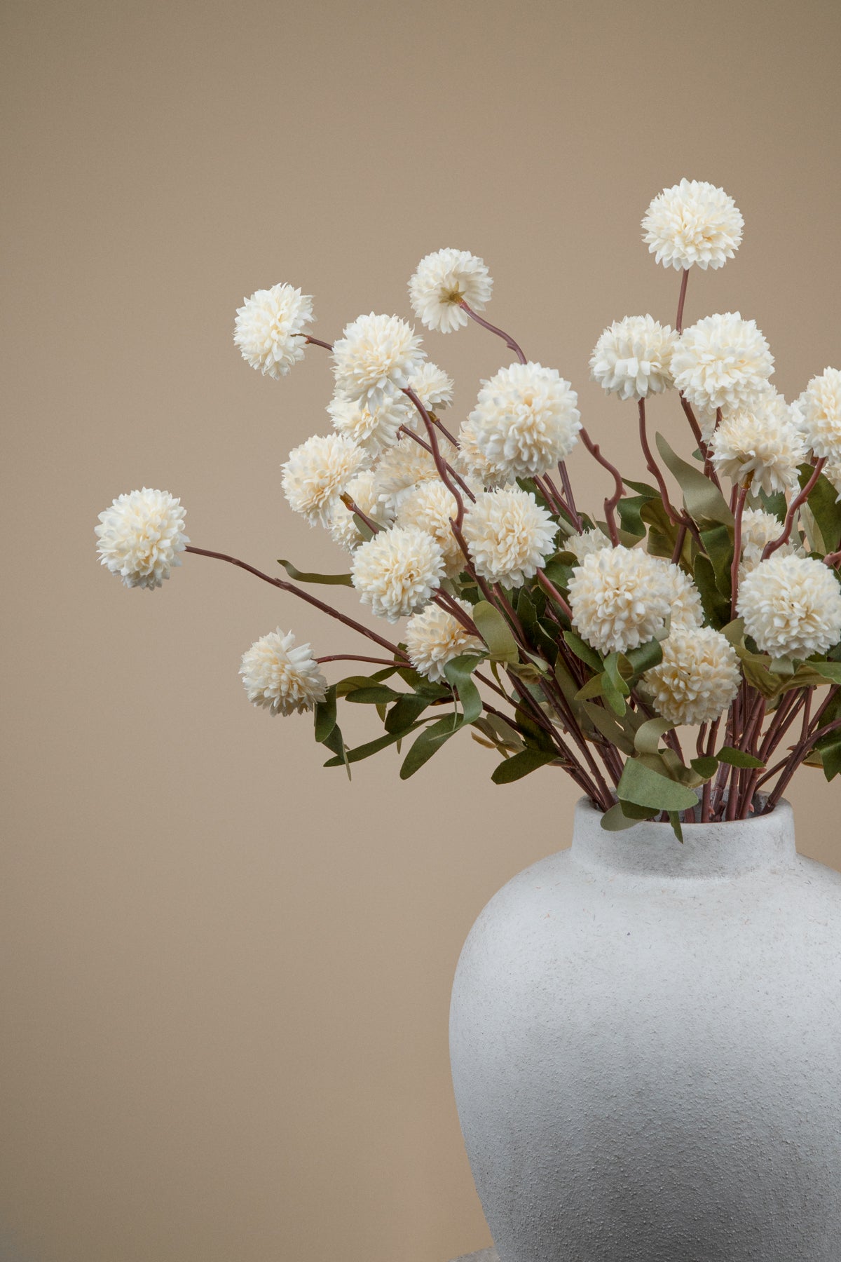 White pom-pom artificial flowers with green leaves arranged in a textured light gray ceramic vase against a solid beige background.