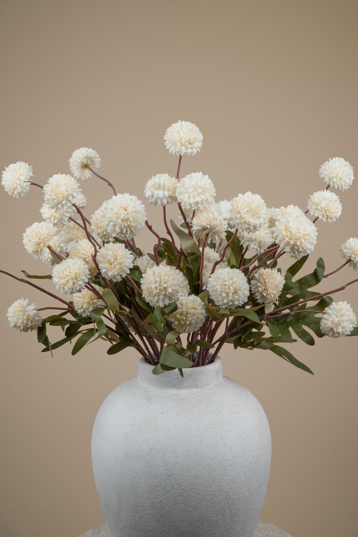 White pom-pom dahlia artificial flowers with green leaves arranged in a matte white ceramic vase against a solid beige background.