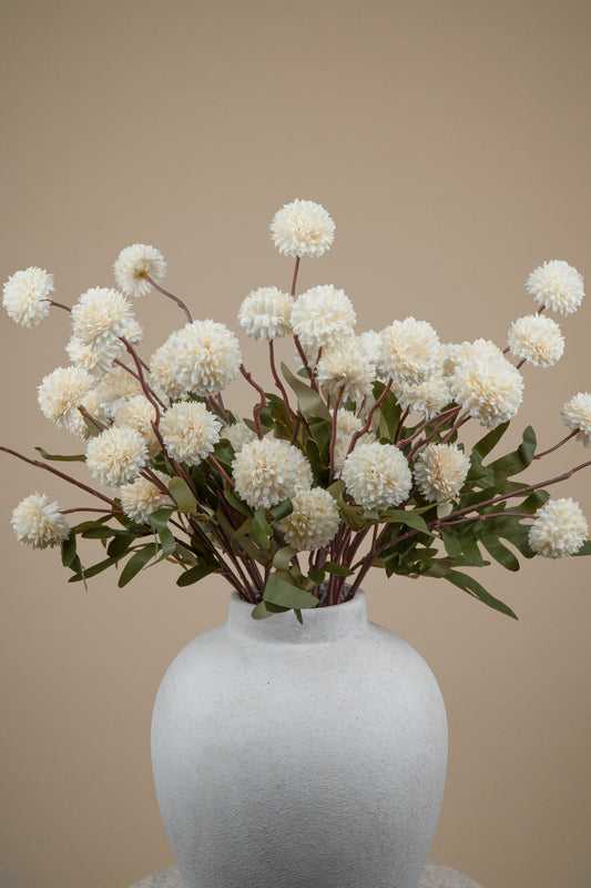 White pom-pom dahlia artificial flowers with green leaves arranged in a matte white ceramic vase against a solid beige background.